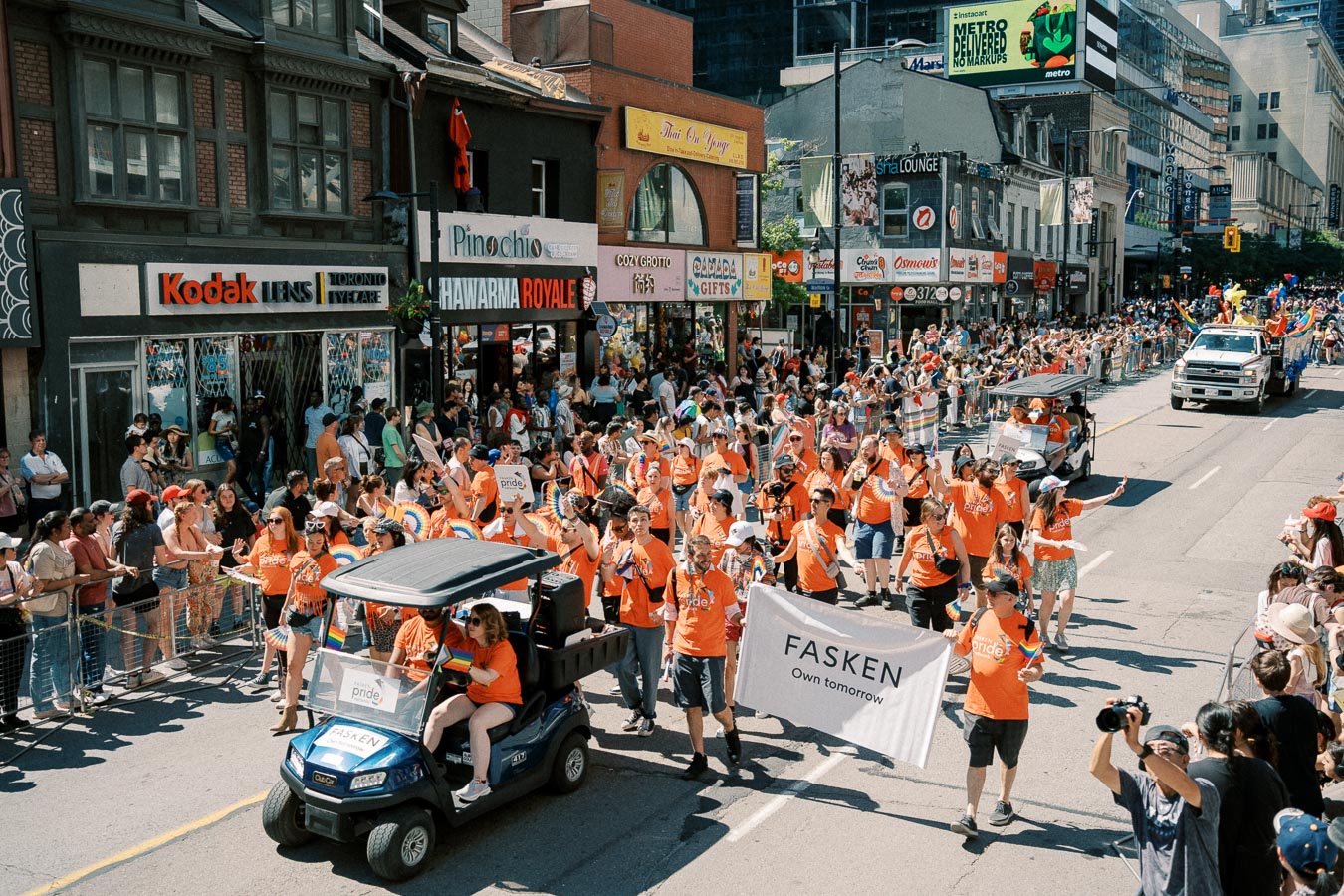 Participants in orange shirts march in a lively street parade, holding a 'FASKEN Own tomorrow' banner. The event is well-attended, with spectators lining the street and various shops in the background.