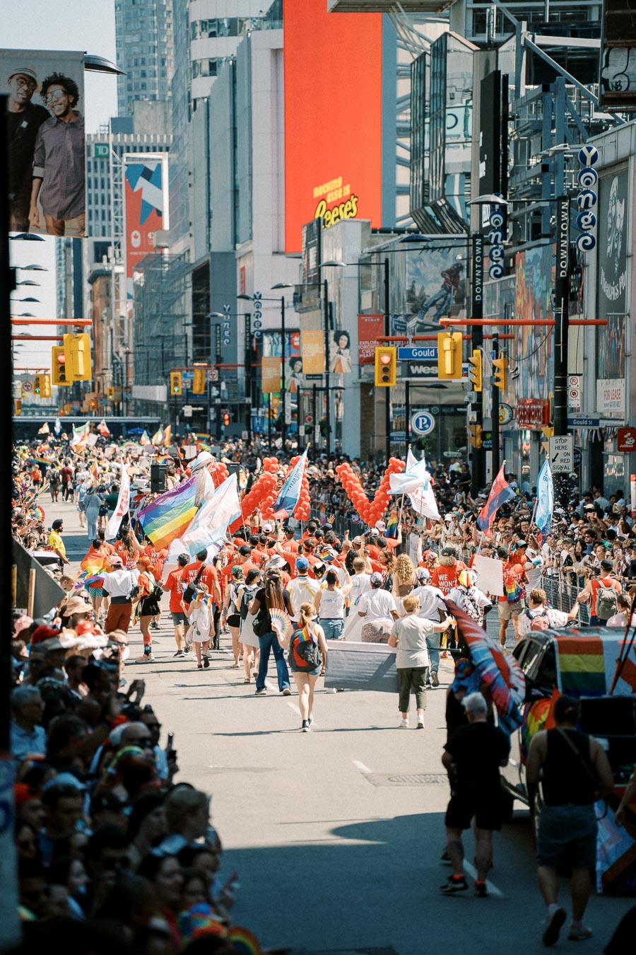 A lively parade takes place on a bustling city street, featuring participants with rainbow flags and balloons, watched by a large crowd. The scene is set against a backdrop of tall buildings and vibrant advertisements.