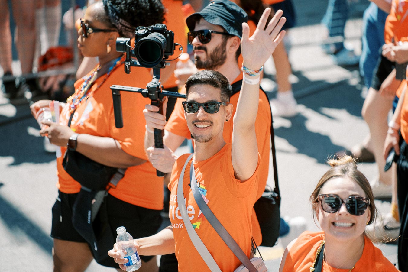 Participants in a parade wearing orange shirts, with one person waving and another filming with a camera on a stabilizer.