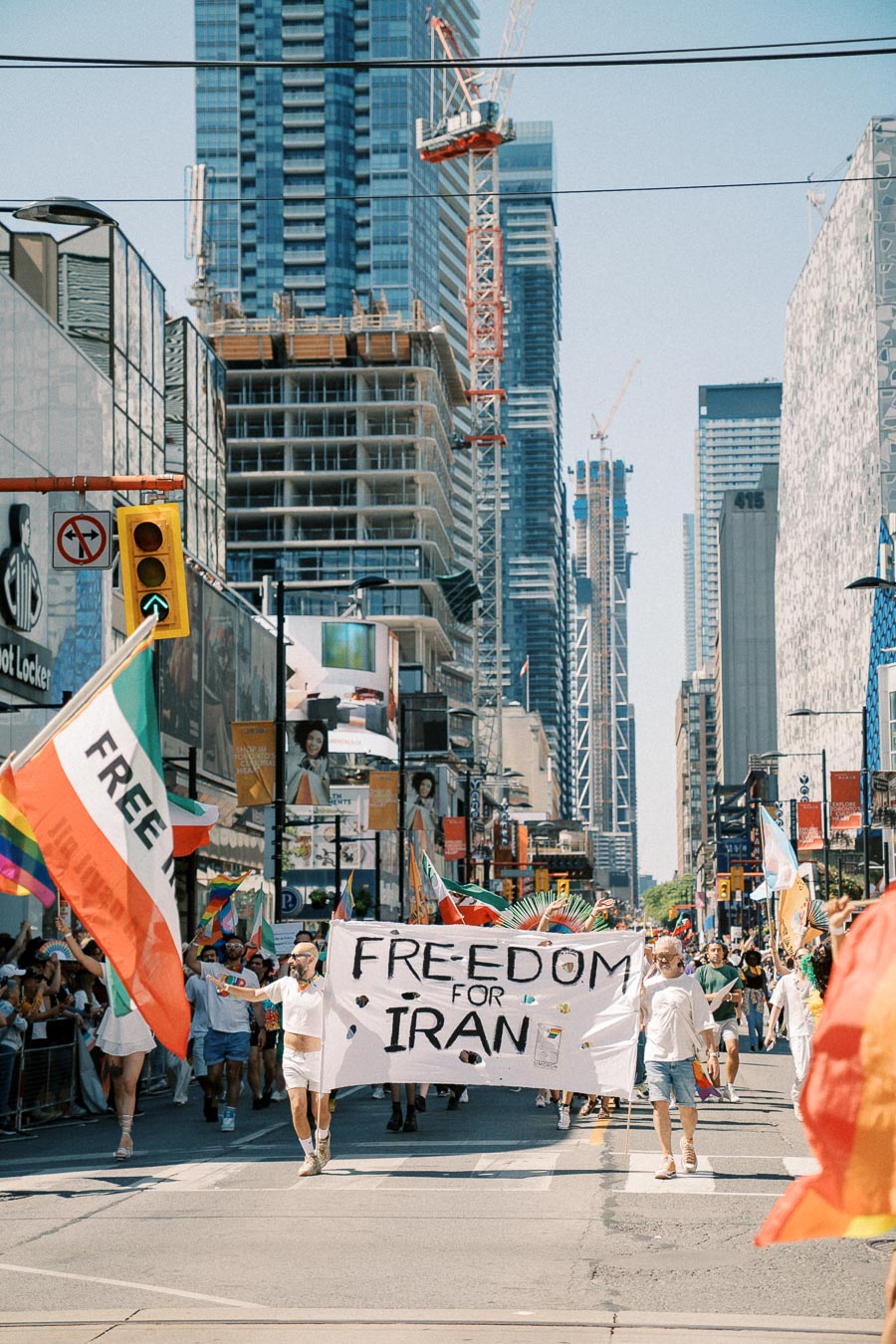 Protesters march in an urban area holding a 'Freedom for Iran' banner, surrounded by tall buildings and construction cranes.