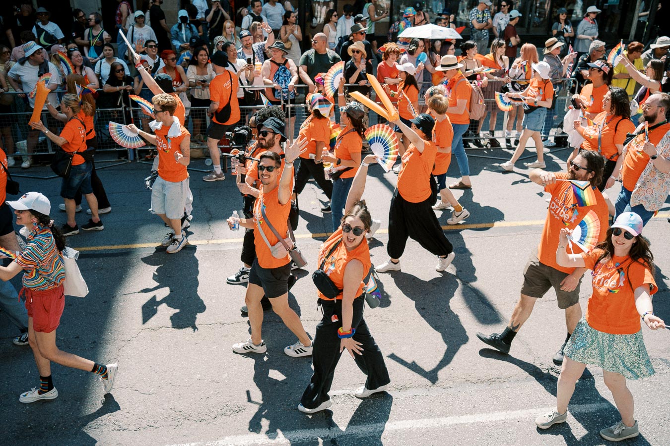 Participants in a pride parade wearing orange shirts and carrying rainbow-themed accessories, while a crowd watches from the sidelines.