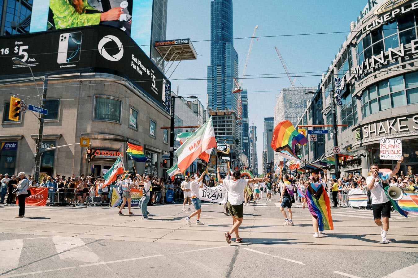 A lively parade takes place in a bustling city intersection, featuring participants waving rainbow and Iranian flags. The scene is set against a backdrop of tall buildings and digital billboards, with a diverse crowd lining the streets.