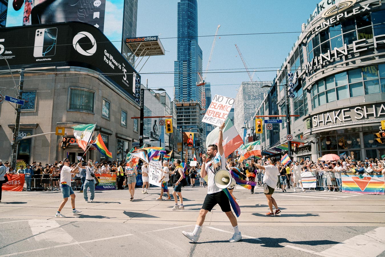 A lively parade takes place at a busy city intersection, with participants carrying flags and signs advocating for freedom and rights. The event is surrounded by a large crowd and tall buildings, creating a festive atmosphere.