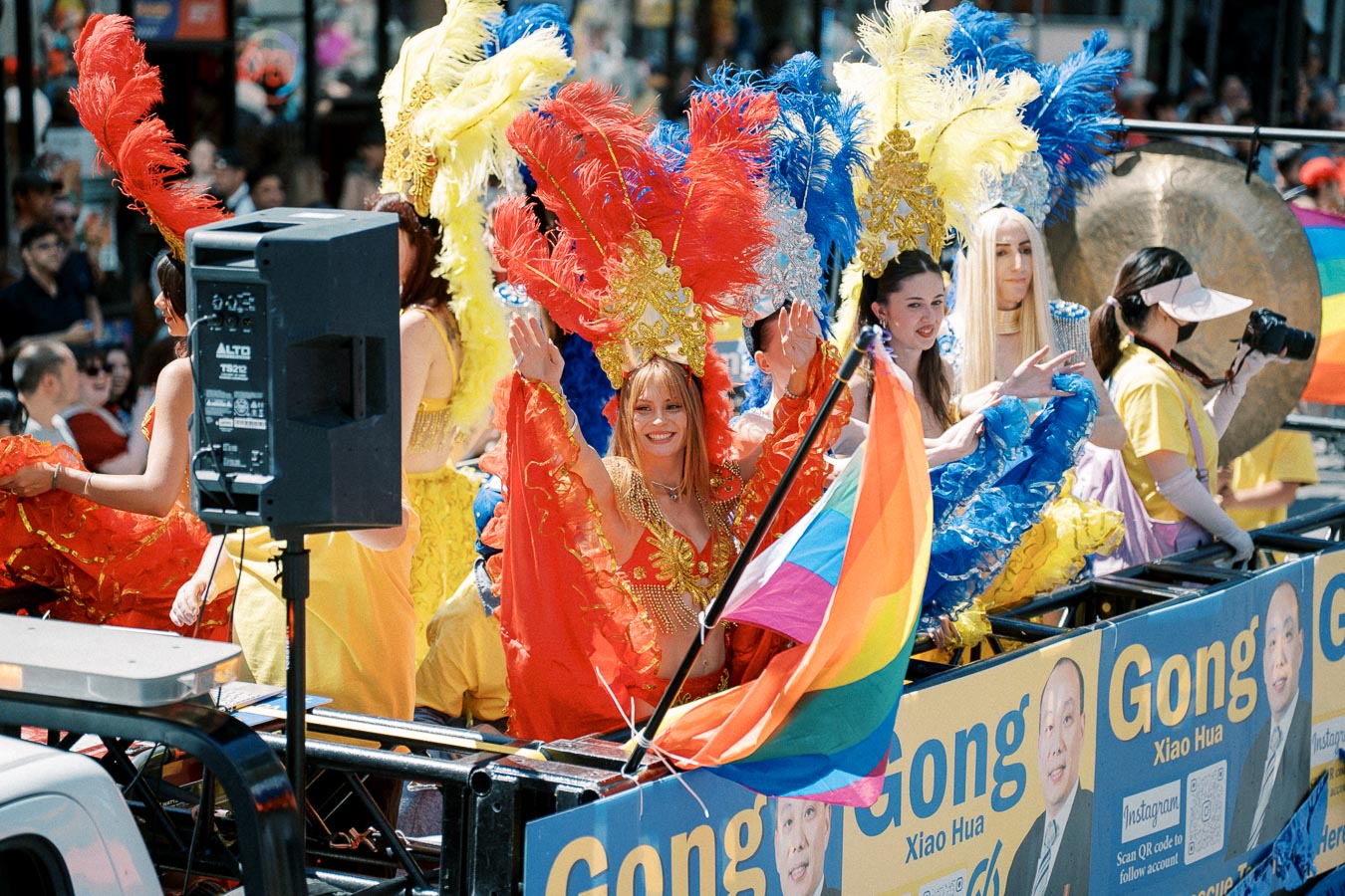 Participants in colorful costumes and feathered headdresses wave from a parade float, displaying a rainbow flag.