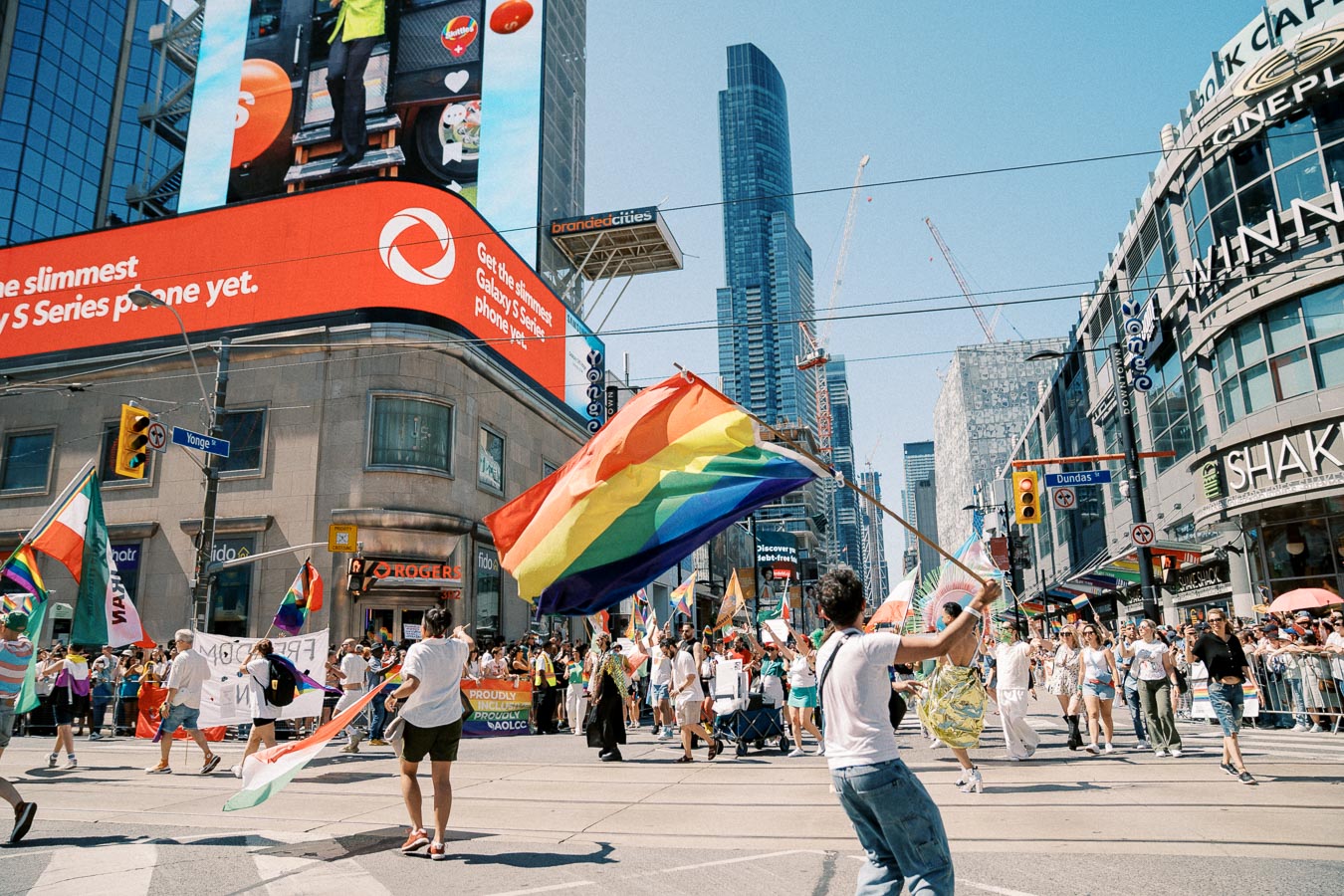 A lively pride parade takes place in a bustling city intersection, with participants waving rainbow flags and banners. The scene is set against a backdrop of tall buildings and digital billboards, celebrating diversity and inclusion.