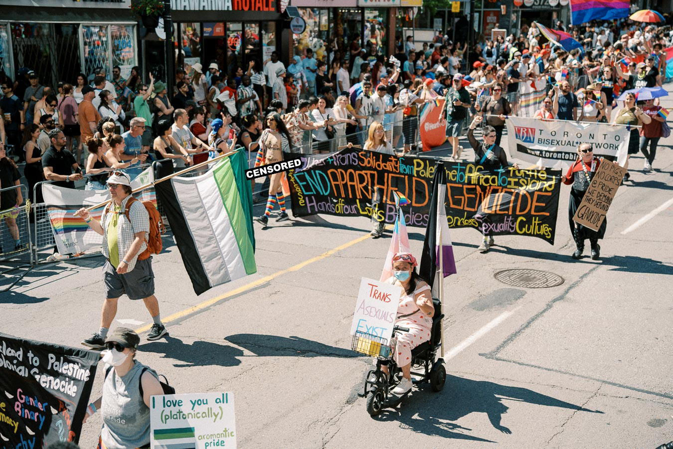 Participants in a parade hold flags and signs advocating for LGBTQ+ rights and social justice, with a large crowd watching from the sidelines.