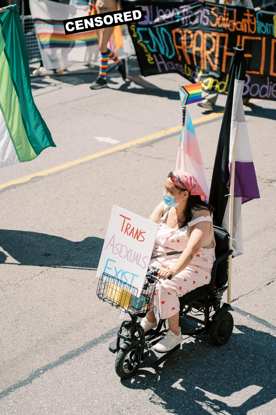 A participant in a pride parade holds a sign reading 'Trans Asexuals Exist' while riding a mobility scooter, surrounded by pride flags.
