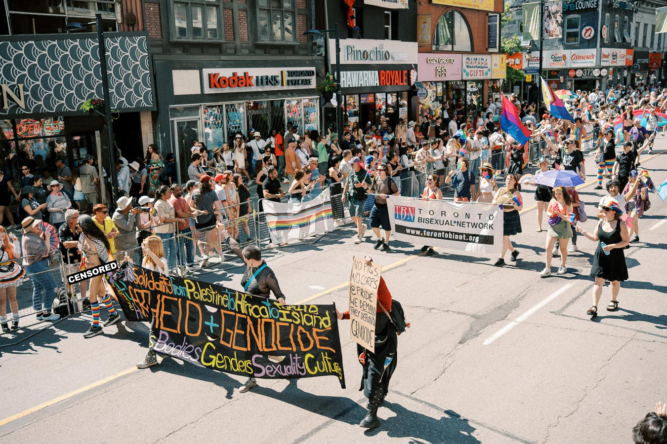 Participants in a pride parade march down a city street, holding banners and flags, including one from the Toronto Bisexual Network, while spectators watch from the sidelines.