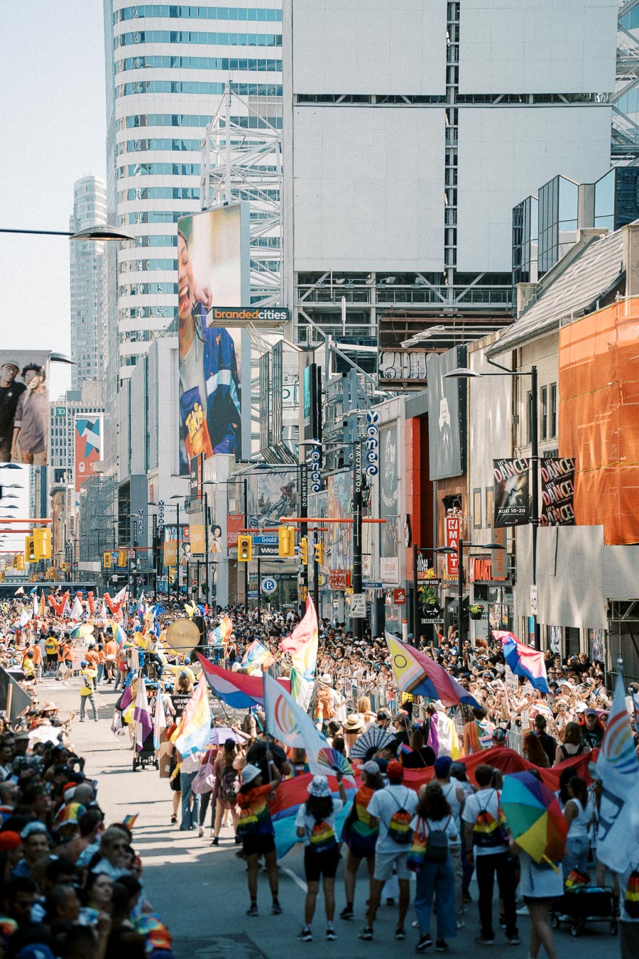 A lively parade featuring rainbow flags and a large crowd takes place on a bustling city street, surrounded by skyscrapers and billboards.