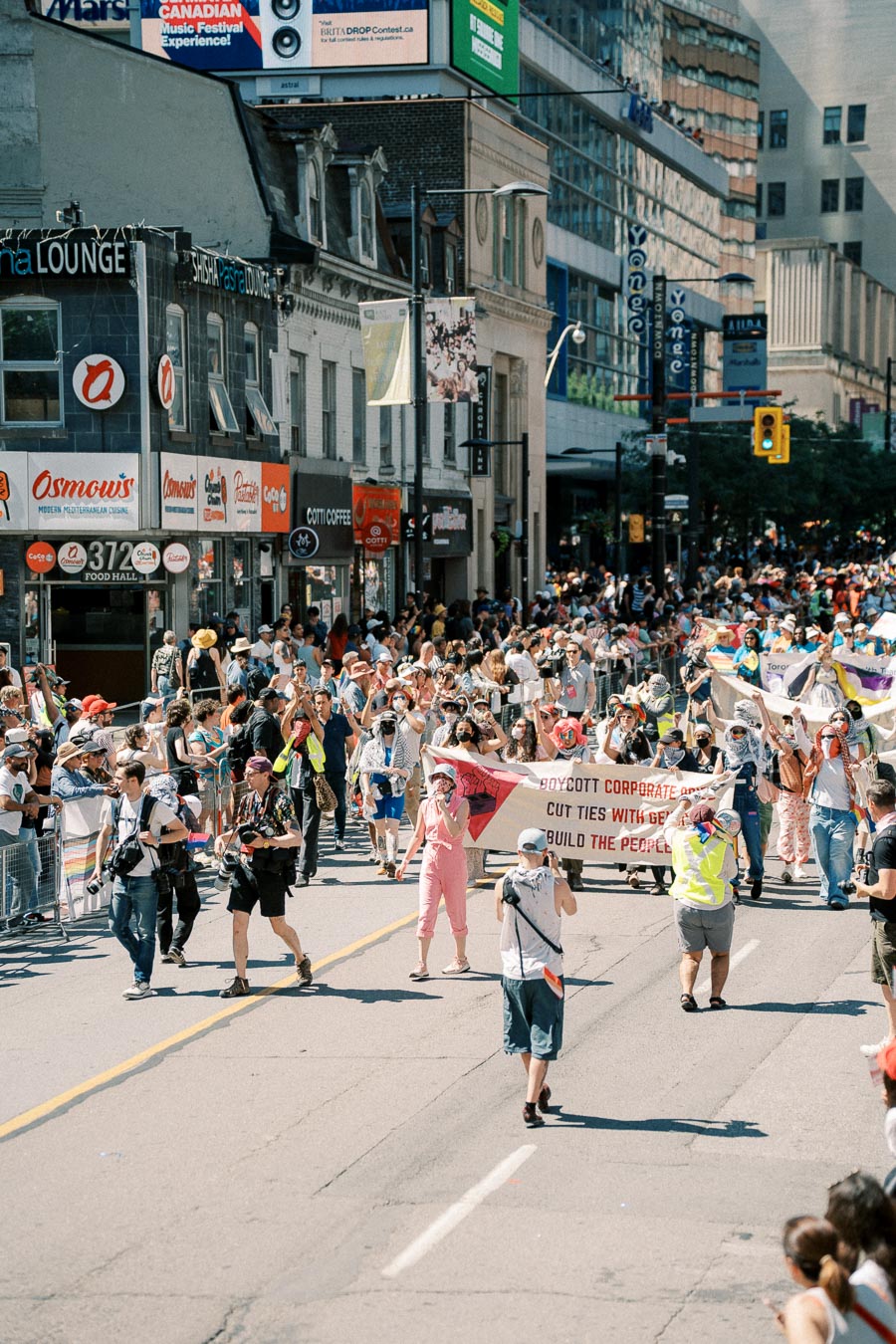 A vibrant street parade with participants holding banners, surrounded by a bustling crowd and city buildings.