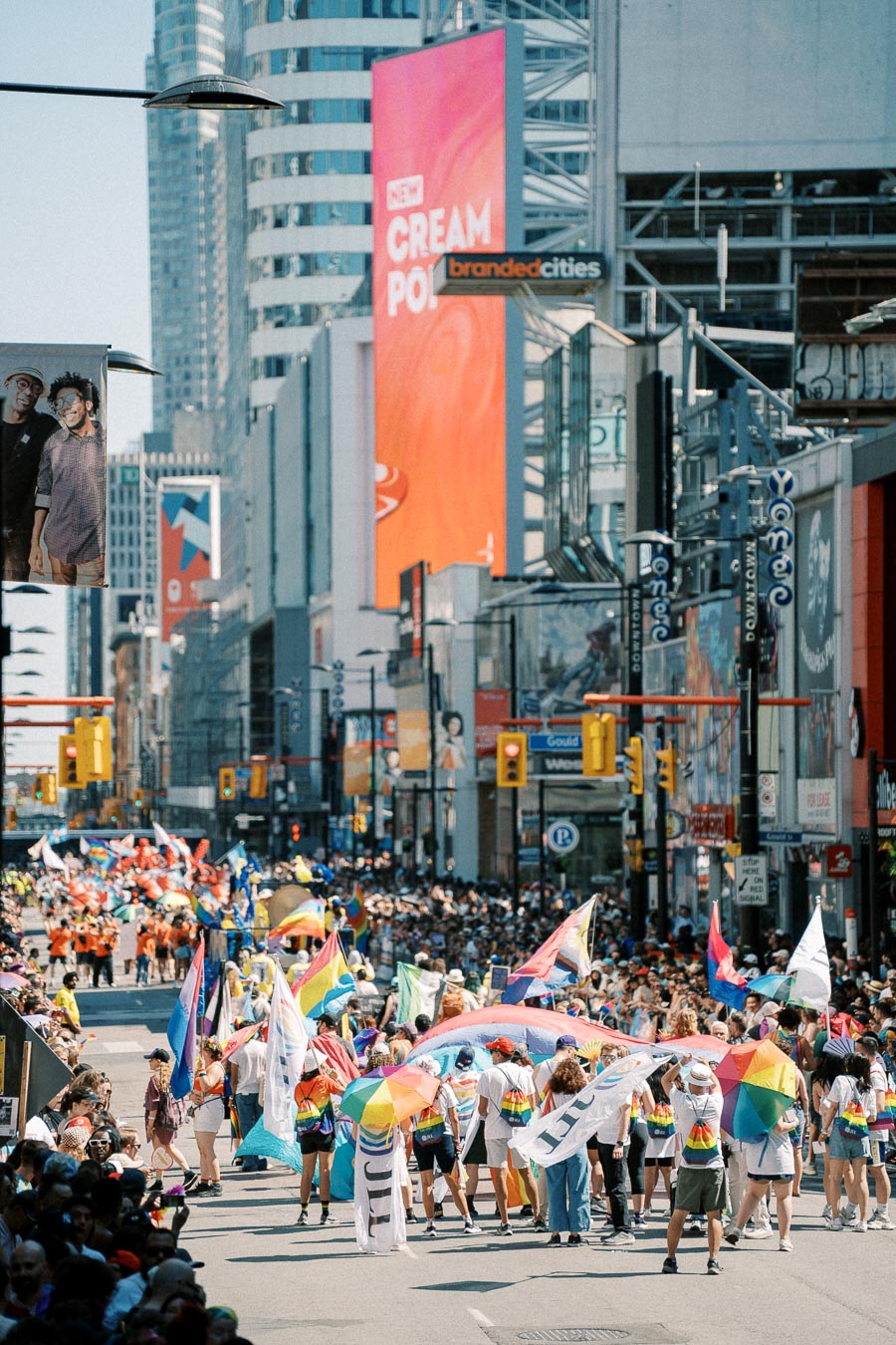 A lively parade featuring rainbow flags and umbrellas takes place on a bustling city street, surrounded by skyscrapers and colorful billboards.