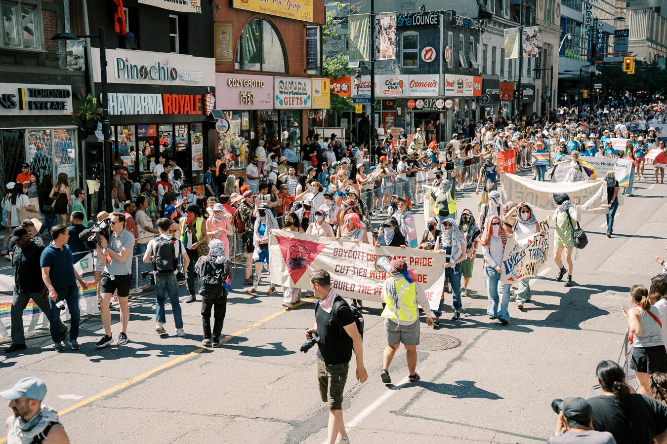 A vibrant parade takes place on a city street, with participants holding banners and wearing colorful attire, while spectators line the sidewalks in front of various shops.