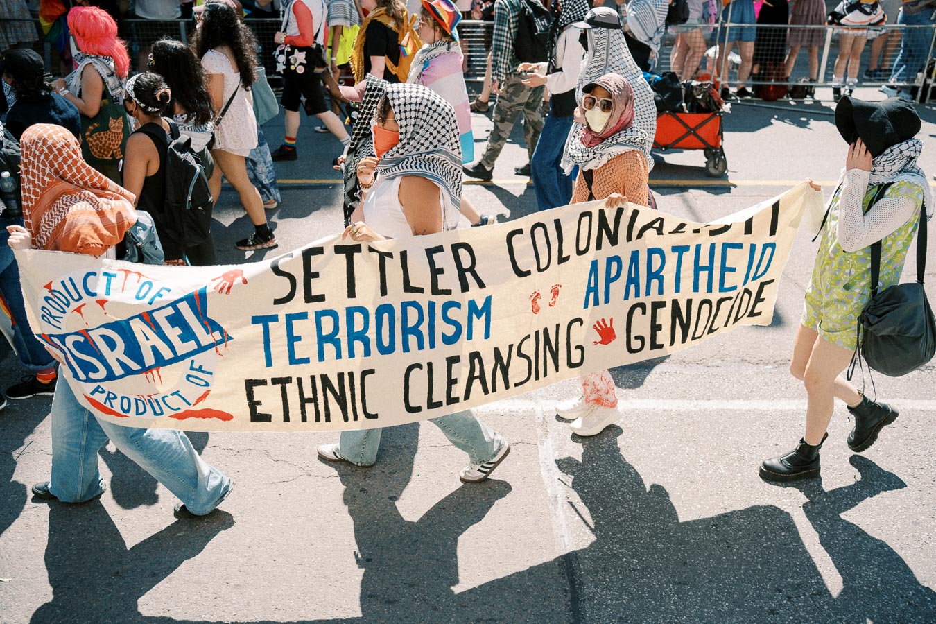 Protesters march holding a banner with political messages related to Israel, including terms like "settler colonialism," "terrorism," "apartheid," "ethnic cleansing," and "genocide." The banner also features a logo with "Product of Israel." Participants are wearing headscarves and masks.