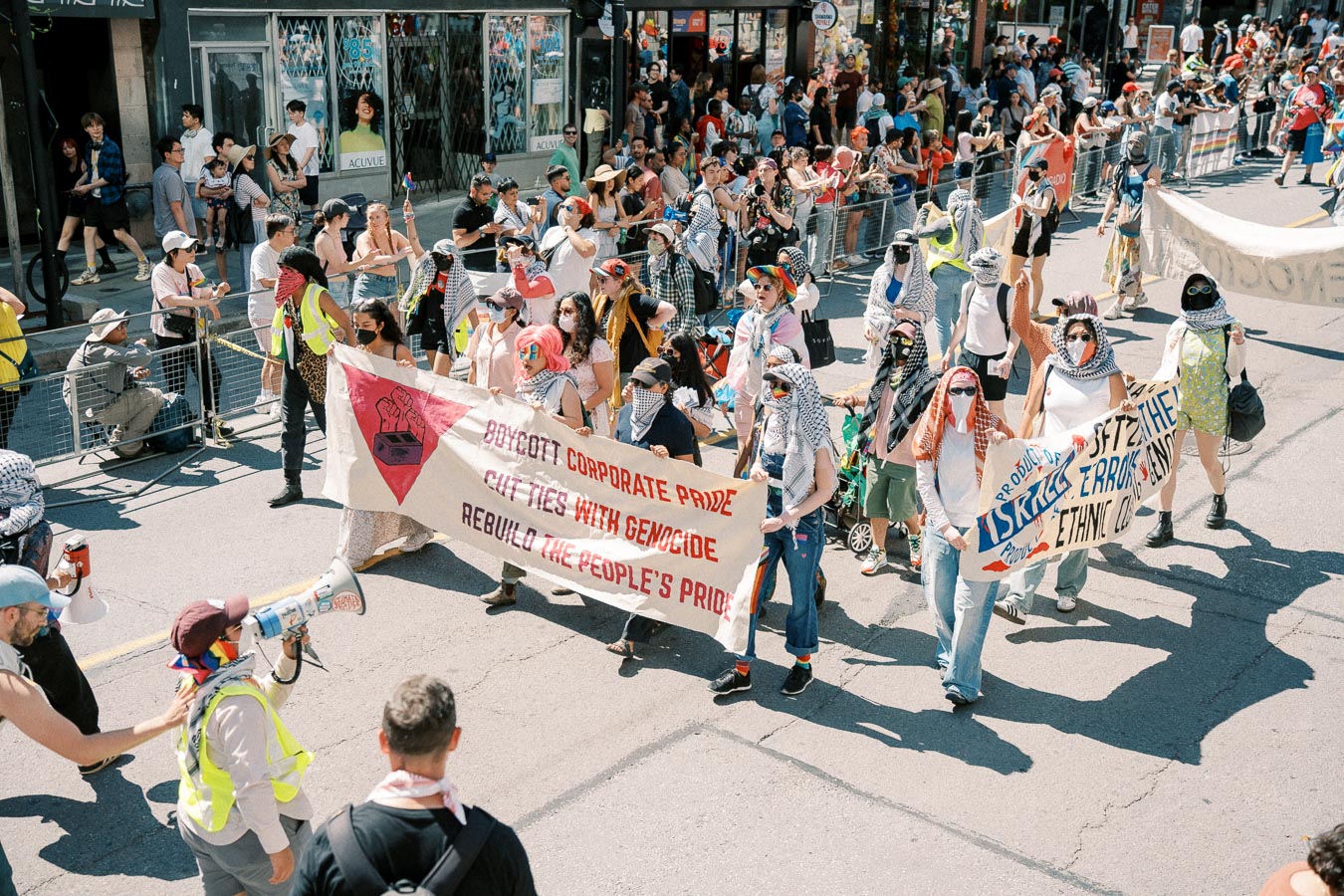 Protesters march down a city street holding banners with messages advocating for boycotting corporate pride and cutting ties with genocide. The crowd is diverse, with many wearing masks and scarves, and spectators line the sidewalks.