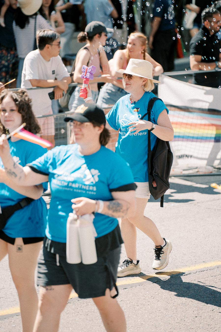 Participants in a parade wearing blue shirts, holding rainbow flags and a bubble blower, with a crowd watching in the background.