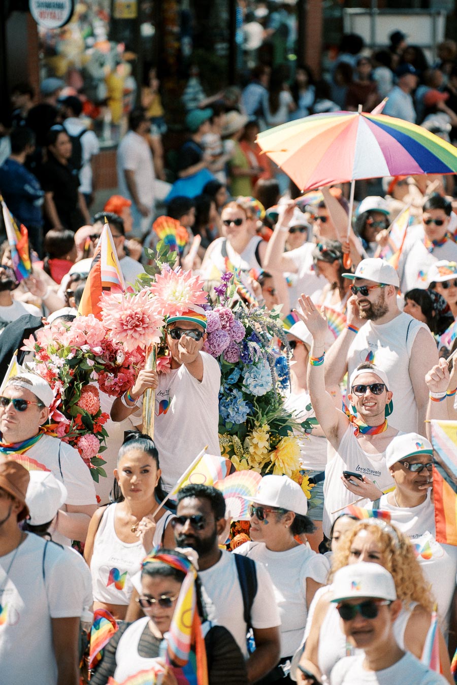 A lively pride parade with participants wearing white shirts and rainbow-themed accessories, surrounded by colorful flowers and a rainbow umbrella.