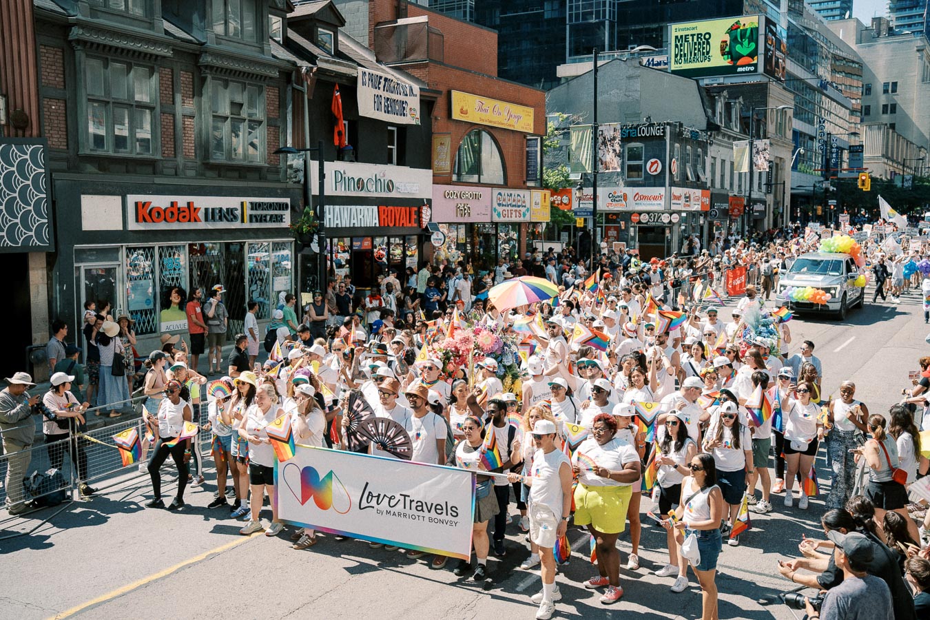 Participants in a pride parade march down a city street, waving rainbow flags and carrying a banner. The street is lined with shops and spectators, creating a festive atmosphere.