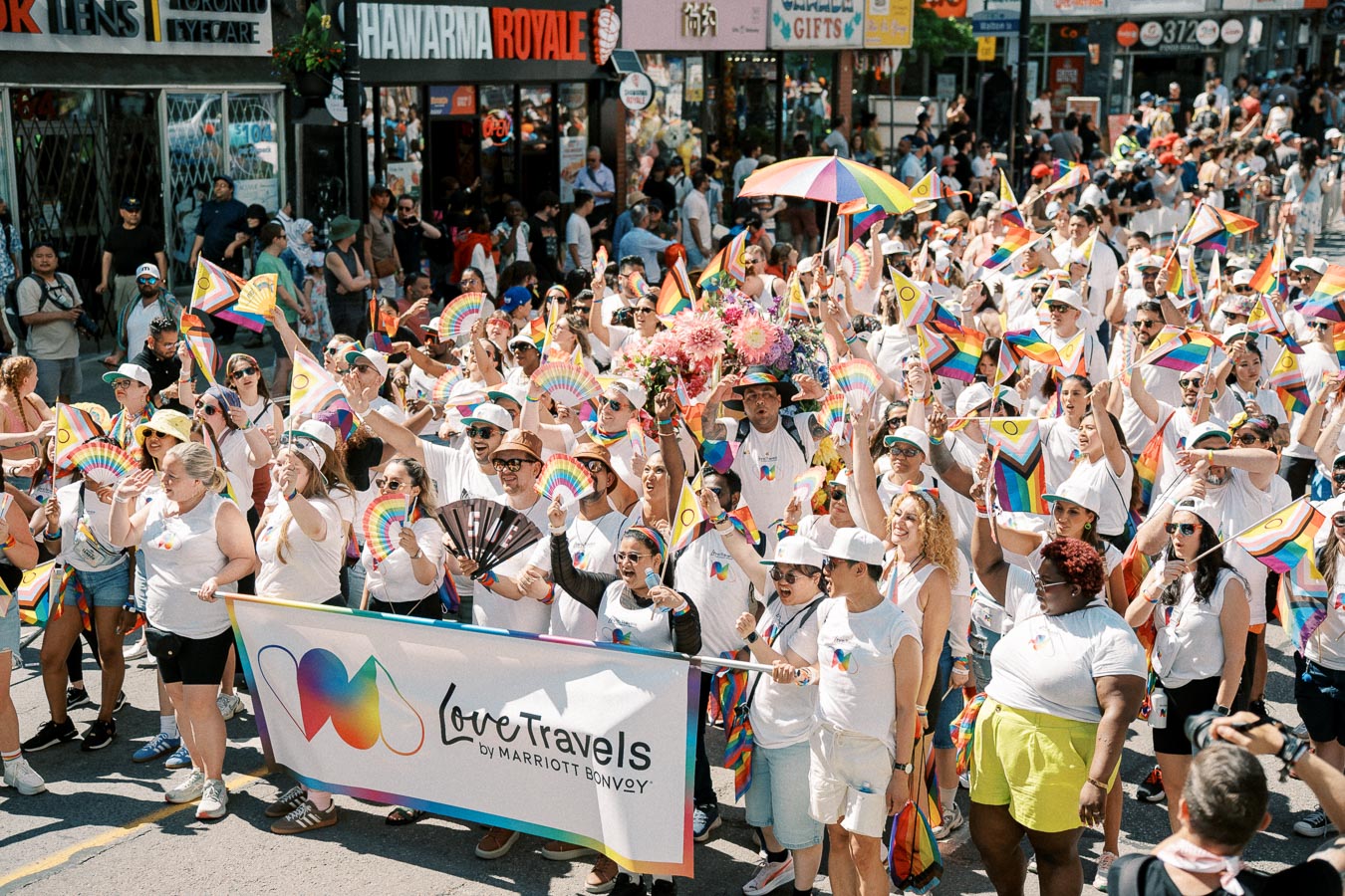 Participants in a Pride parade celebrate with rainbow flags and fans, marching under the banner 'Love Travels by Marriott Bonvoy'.