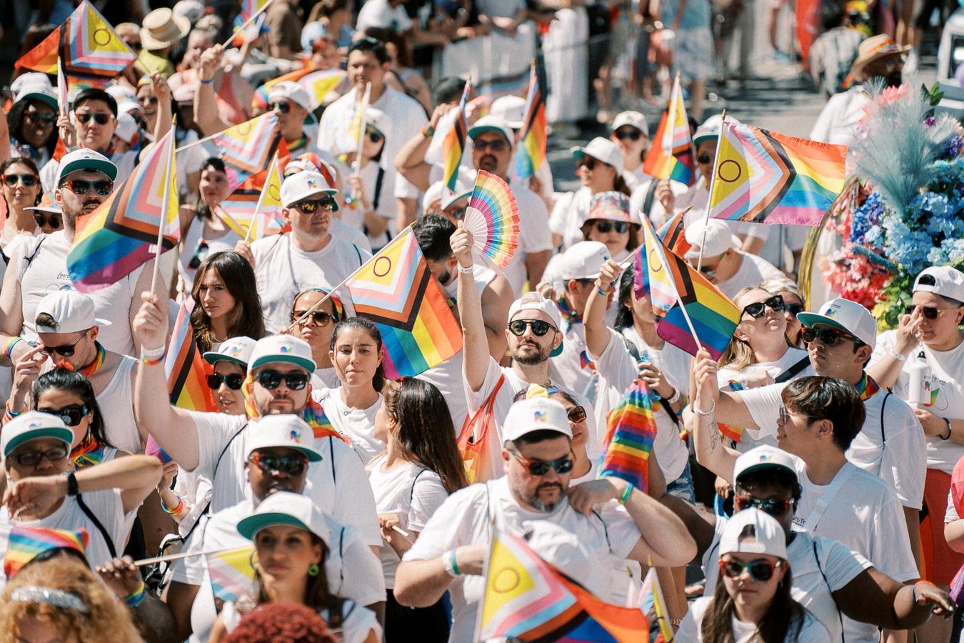 Participants in a pride parade celebrate diversity and inclusion, waving rainbow flags and wearing white attire.