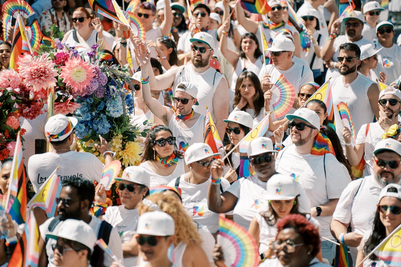 Participants celebrate at a pride parade, adorned with rainbow flags and accessories, surrounded by vibrant flowers.