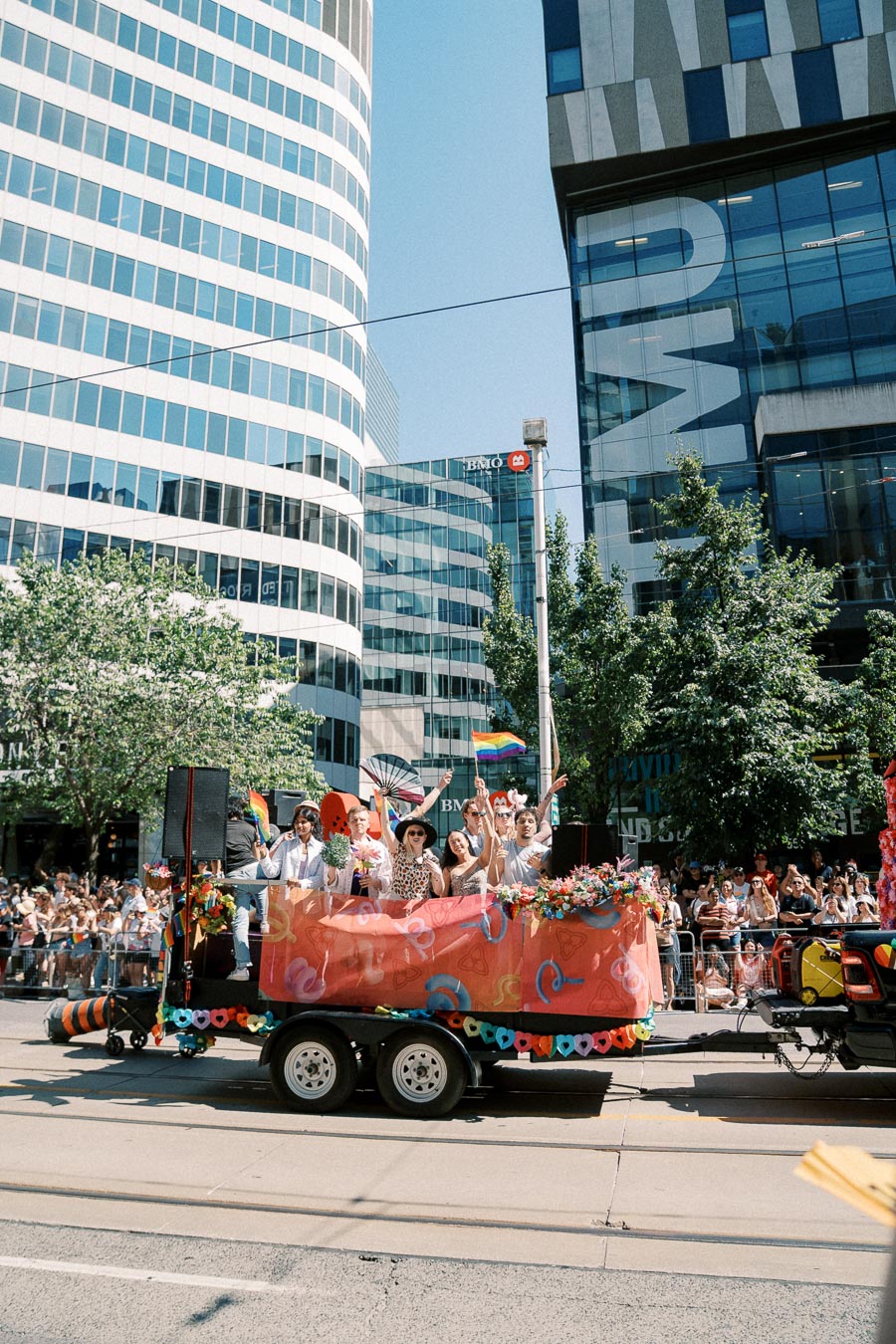 A vibrant float adorned with rainbow flags and decorations moves through a city street during a pride parade, with modern office buildings in the background.