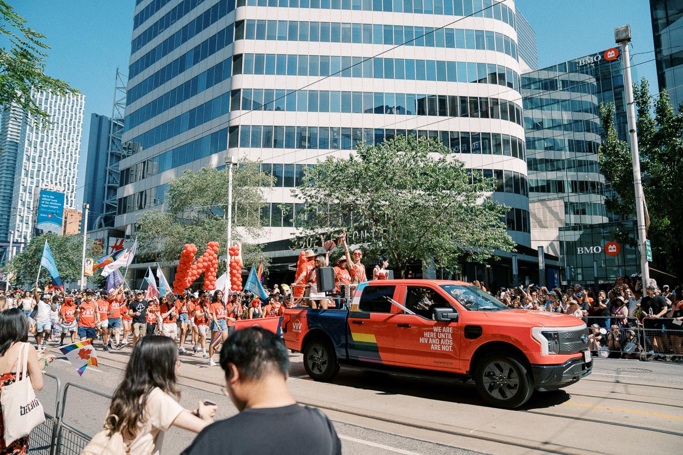 A lively parade scene featuring a red truck with rainbow decorations and participants marching, surrounded by modern city buildings.