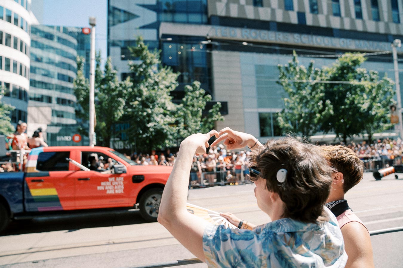 Spectators form a heart shape with their hands at a parade, featuring a colorful truck and a lively crowd in front of the Ted Rogers School of Management.