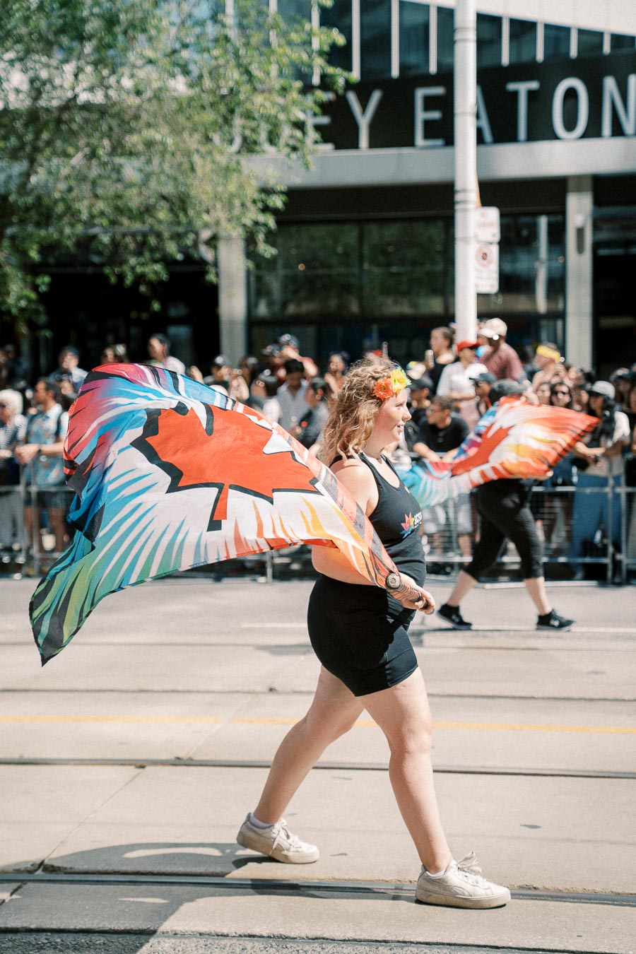 A participant in a parade holds a vibrant flag featuring a maple leaf design, symbolizing Canada. The event takes place on a sunny day with a crowd watching in the background.