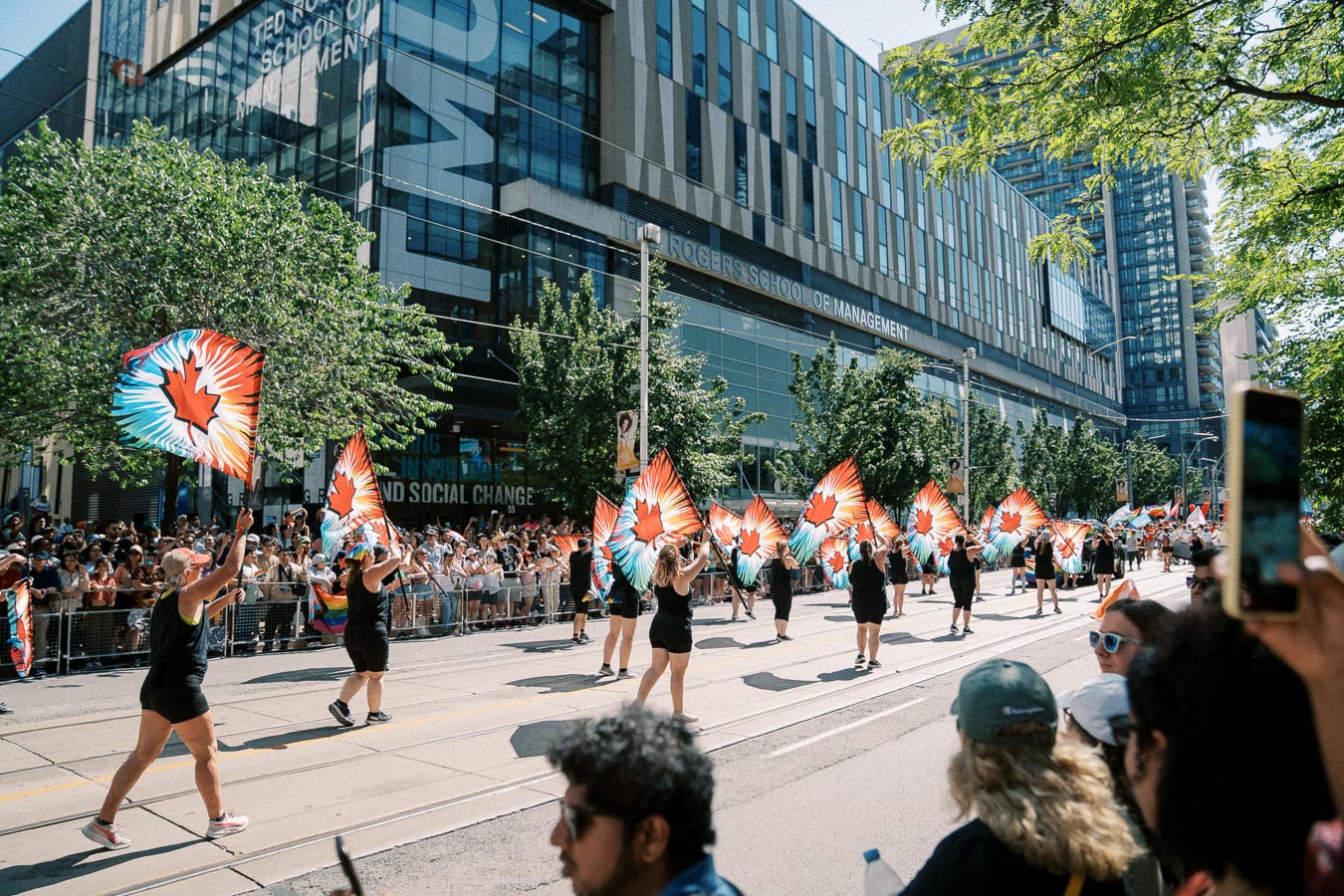 Participants in a parade wave vibrant flags with a maple leaf design, marching past the Ted Rogers School of Management.