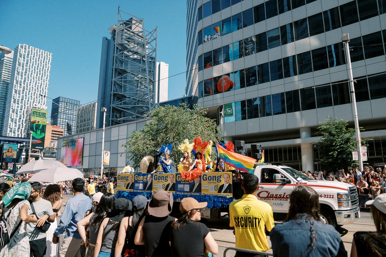 A vibrant parade float featuring people in colorful costumes and rainbow flags moves through a city street, surrounded by a large crowd of spectators.