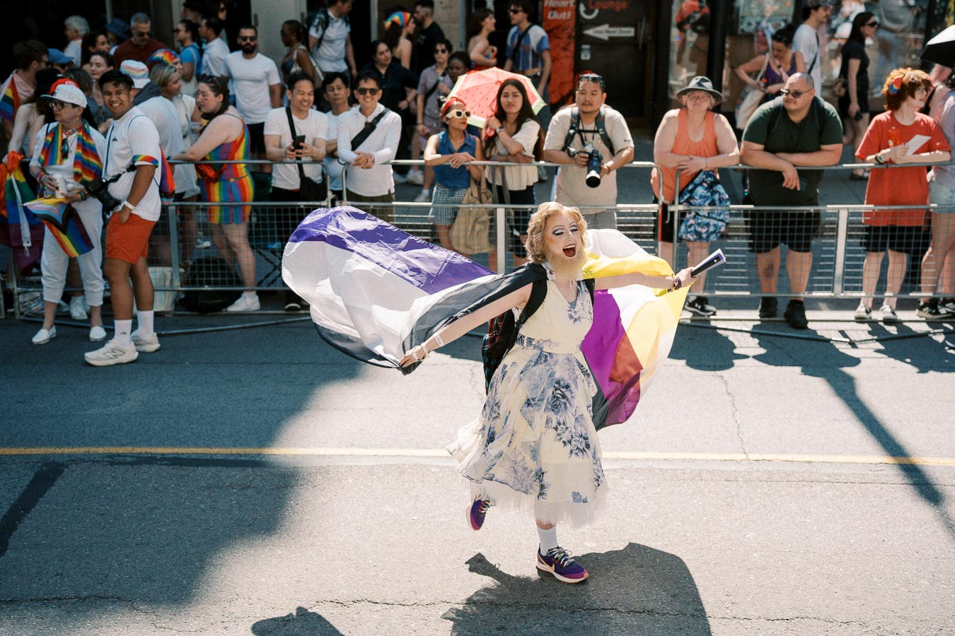 A person joyfully waves a non-binary pride flag during a parade, surrounded by a cheering crowd.