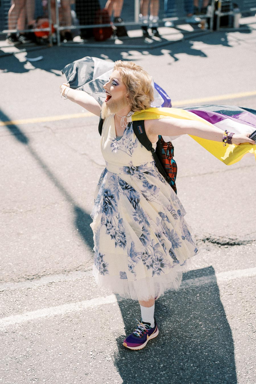A person joyfully participates in a parade, wearing a floral dress and holding a non-binary pride flag. They have a backpack and are wearing purple sneakers.