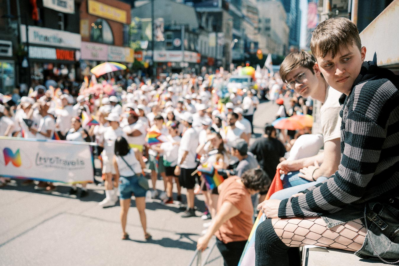 Participants in a pride parade fill the street, while two individuals observe from a ledge.