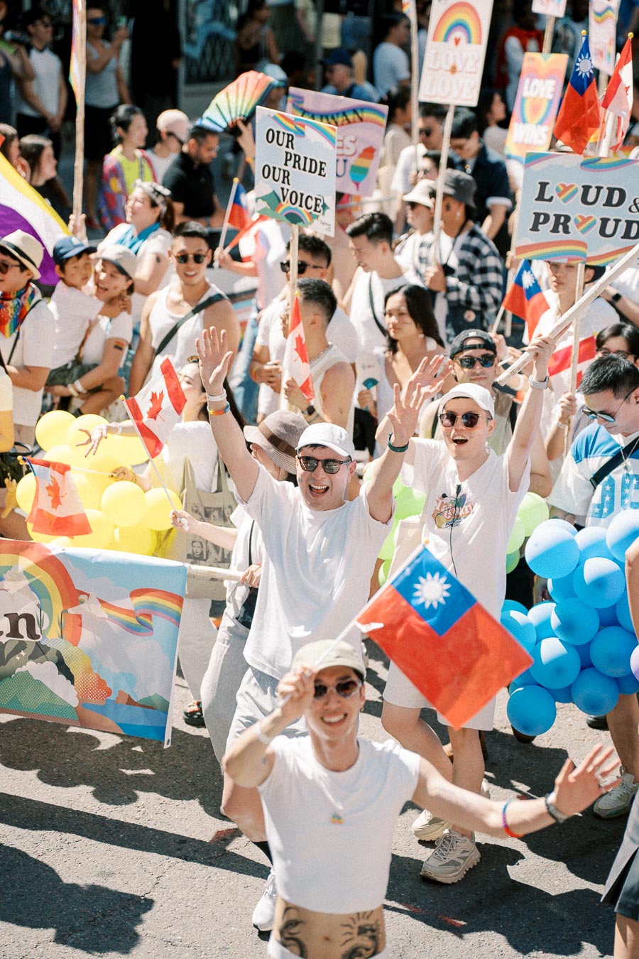 Participants celebrate at a pride parade, waving Canadian and Taiwanese flags, and holding rainbow-themed signs and balloons.