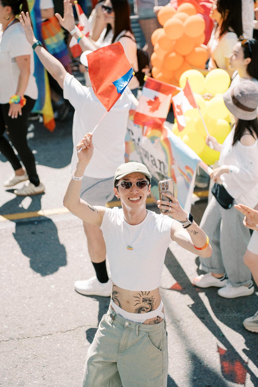 A joyful participant at a parade holds a Taiwanese flag and takes a selfie, surrounded by others with Canadian flags and vibrant balloons.