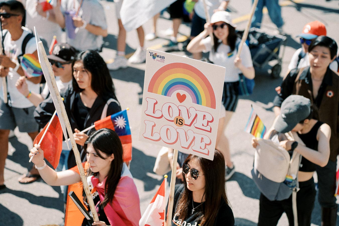 Participants at a pride parade hold a 'Love is Love' sign and flags from Taiwan and Canada, celebrating diversity and equality.