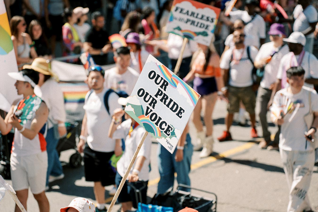 Participants in a pride parade hold colorful signs, including one that reads 'Our Pride Our Voice', celebrating diversity and inclusion.