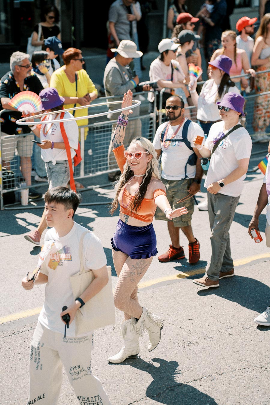Participants in a vibrant pride parade, featuring a woman in orange and purple attire with heart-shaped sunglasses, surrounded by a diverse crowd.