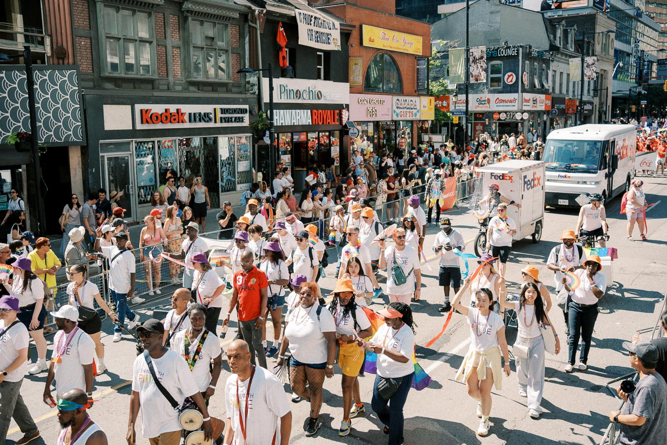 Participants in a lively street parade walk past shops and spectators, showcasing colorful attire and festive spirit.