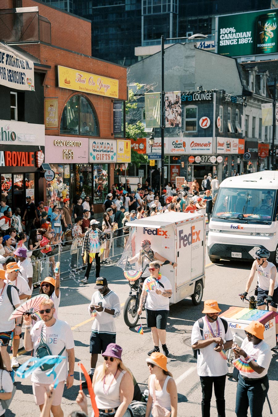A vibrant parade scene with participants in colorful attire, featuring a FedEx vehicle, on a bustling street lined with shops and spectators.
