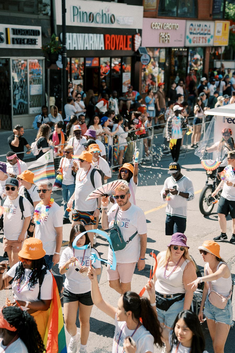 Participants in a pride parade walk down a city street, wearing colorful accessories and shirts with rainbow text, as spectators watch from the sidelines.