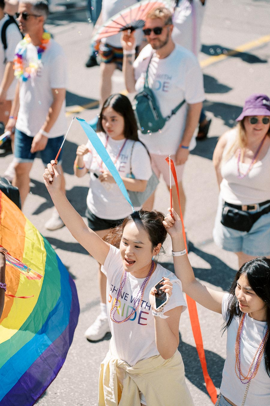 Participants celebrate at a pride parade, wearing shirts with rainbow text and waving colorful ribbons and flags.