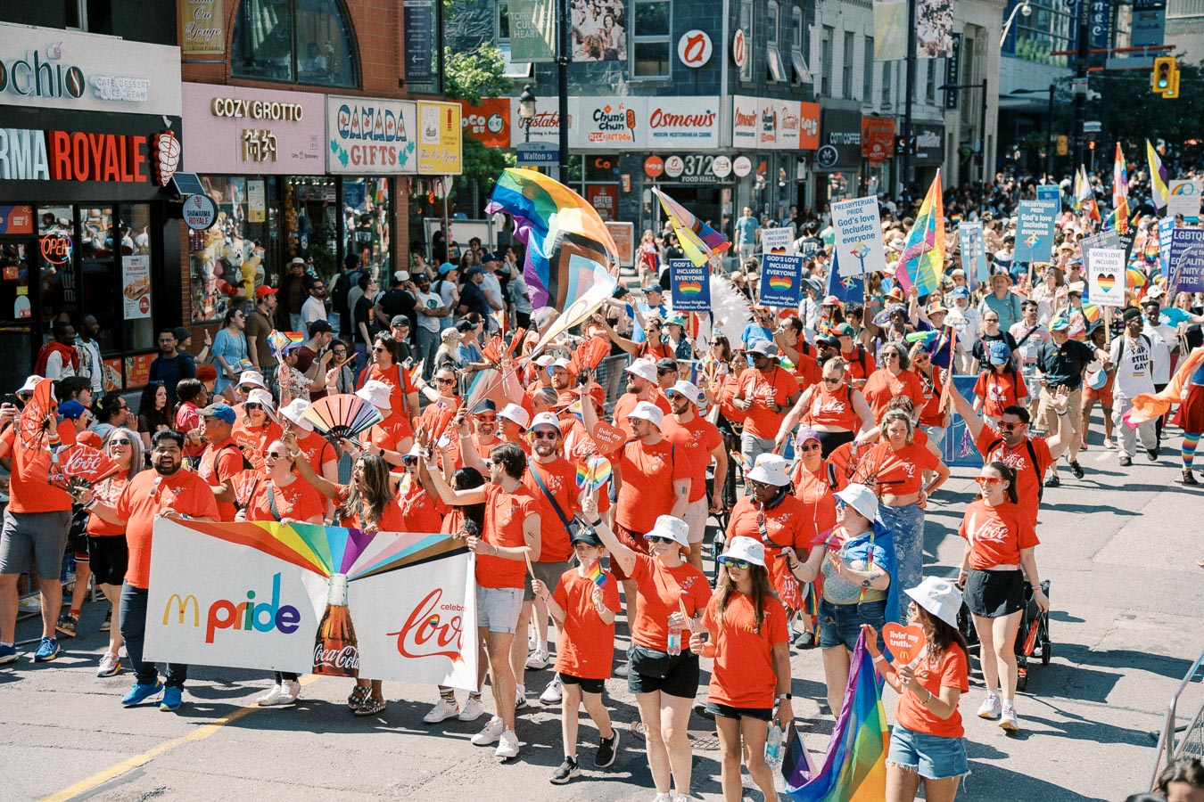 Participants in a Pride parade march down a city street, wearing red shirts and white hats, and carrying rainbow flags and banners. The event is lively and colorful, with many spectators lining the street.