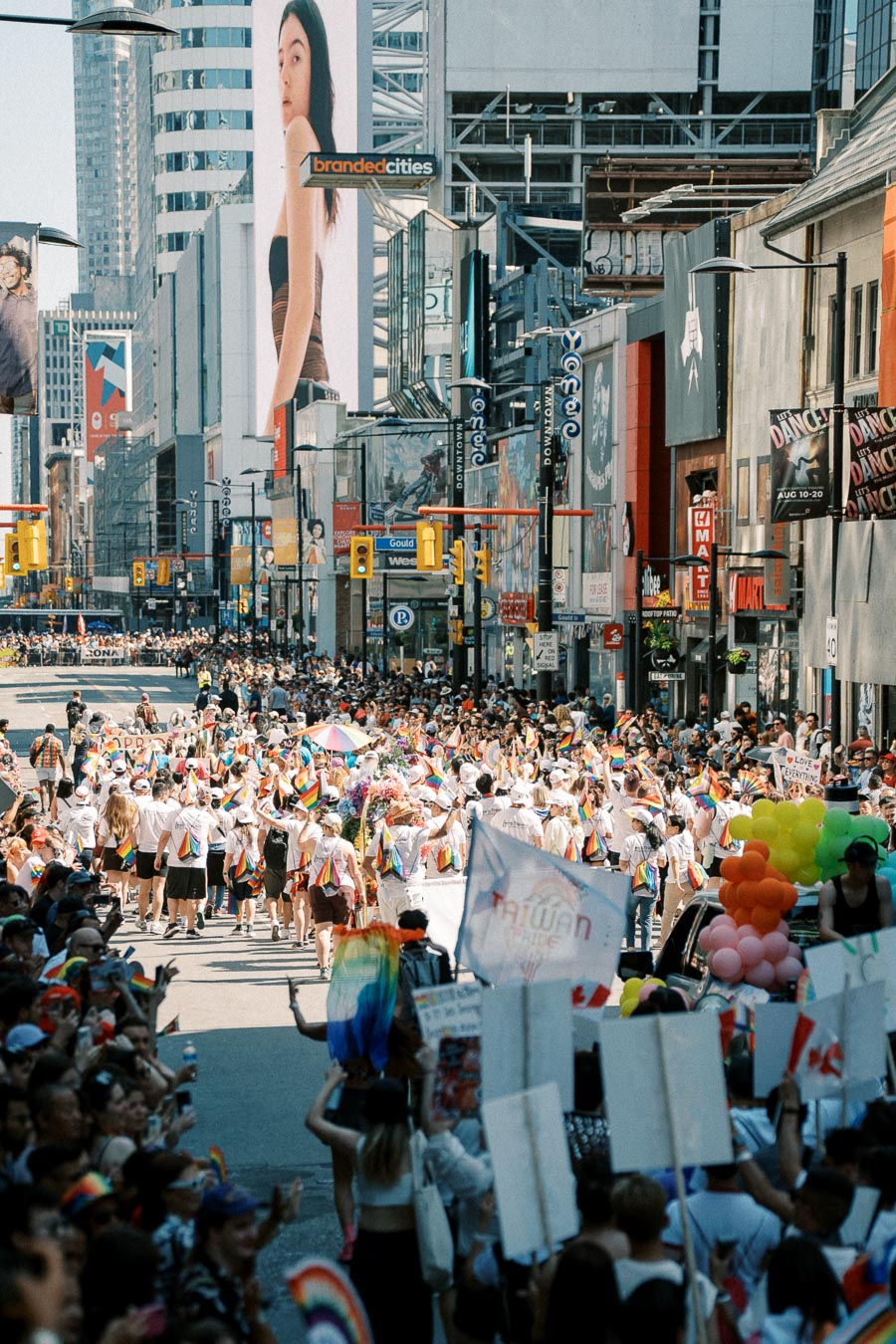 A lively pride parade takes place on a bustling city street, with participants waving rainbow flags and carrying banners. The street is lined with spectators and flanked by tall buildings adorned with large digital billboards.