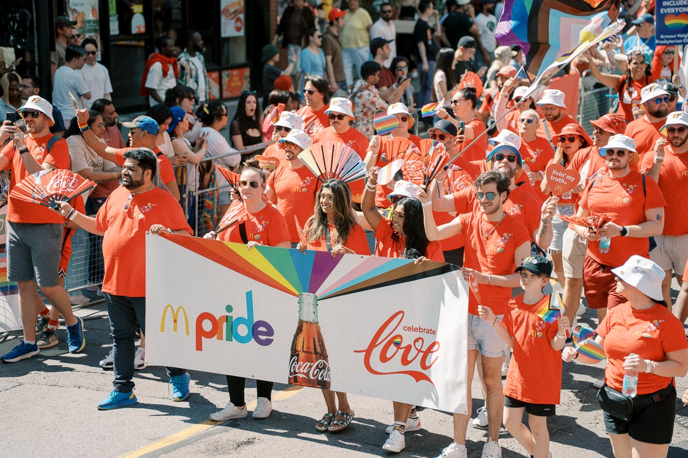Participants in a Pride parade wearing red shirts and holding a banner promoting inclusivity and love.