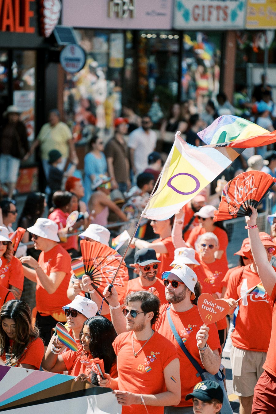 Participants in a pride parade wearing red shirts and white hats, holding rainbow flags and fans.