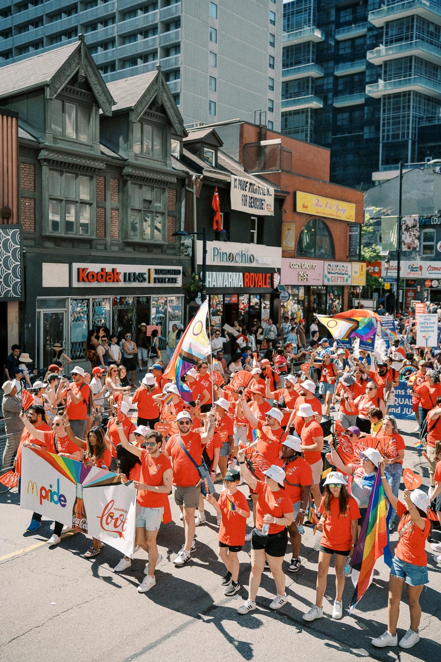 Participants in a pride parade march down a city street, displaying rainbow flags and banners, surrounded by urban buildings and shops.
