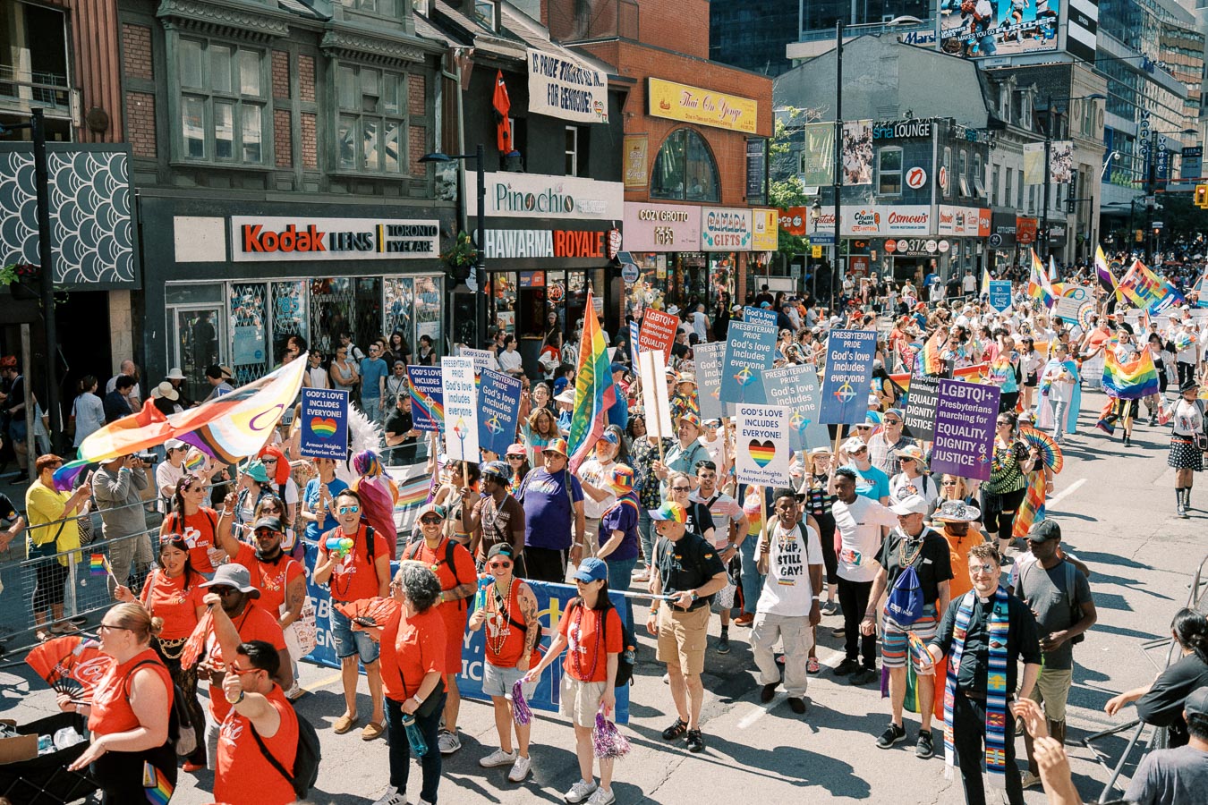 Participants in a pride parade march down a city street, holding signs promoting inclusivity and equality, with rainbow flags and colorful attire.