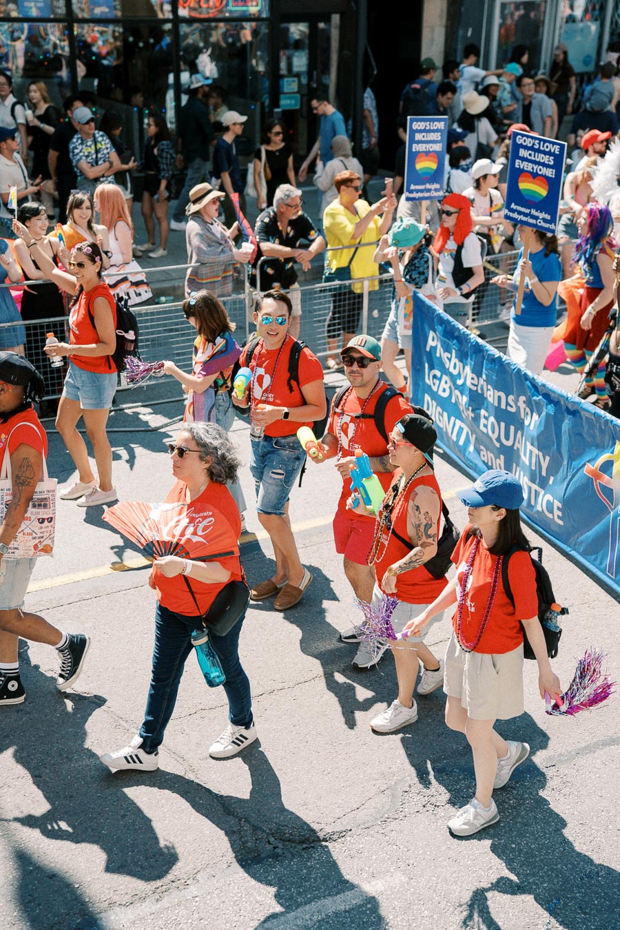 Participants in a pride parade march with signs advocating for LGBTQ+ equality and inclusion, wearing red shirts and carrying colorful accessories.
