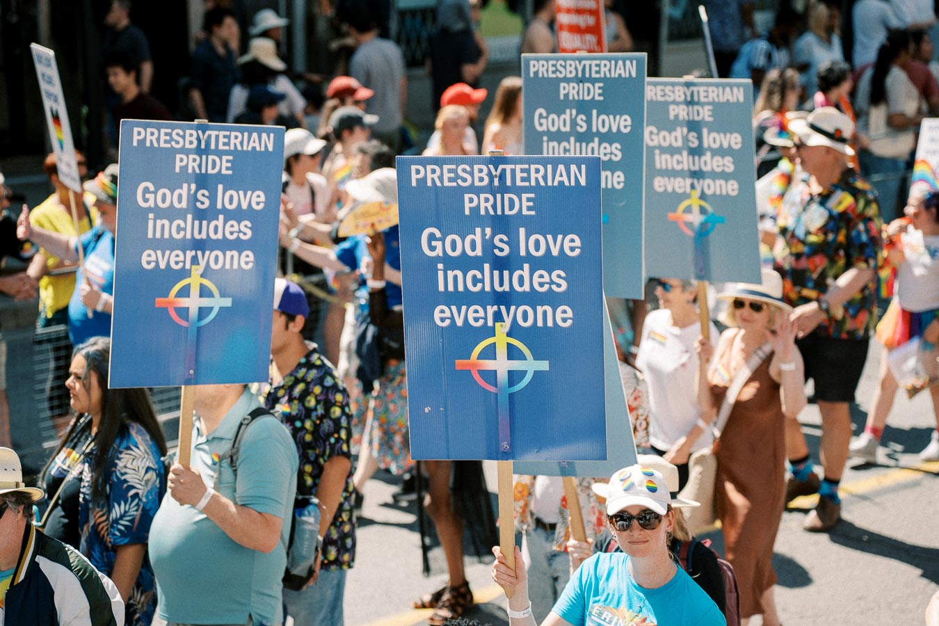 Participants in a parade hold signs reading 'Presbyterian Pride: God's love includes everyone', promoting inclusivity and acceptance.